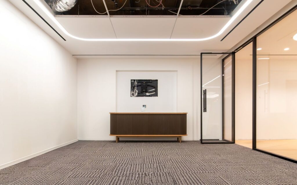 A wide, symmetrical view of a minimalist office or meeting room under construction at the 1100 NY Ave project. The focal point is a dark wood-slatted sideboard with rounded ends and a light-wood top, centered against a white wall beneath a recessed TV mount.
