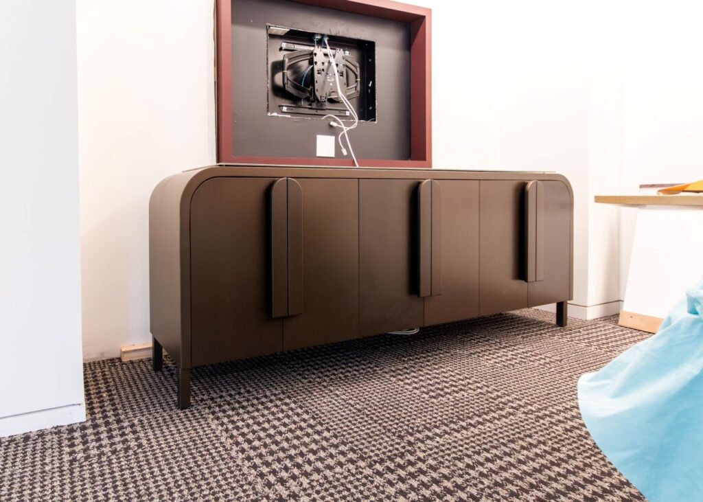 A detailed shot of the dark-bronze metal sideboard from the "1100 NY Ave" project, featuring its distinctive curved silhouette and three vertical pill-shaped door handles. The unit is placed on a brown-and-white checkered carpet, sitting beneath a recessed wall cutout framed in burgundy that contains exposed television mounting hardware and wiring. To the right, the edge of a light-wood table and a blue fabric-covered object are partially visible.