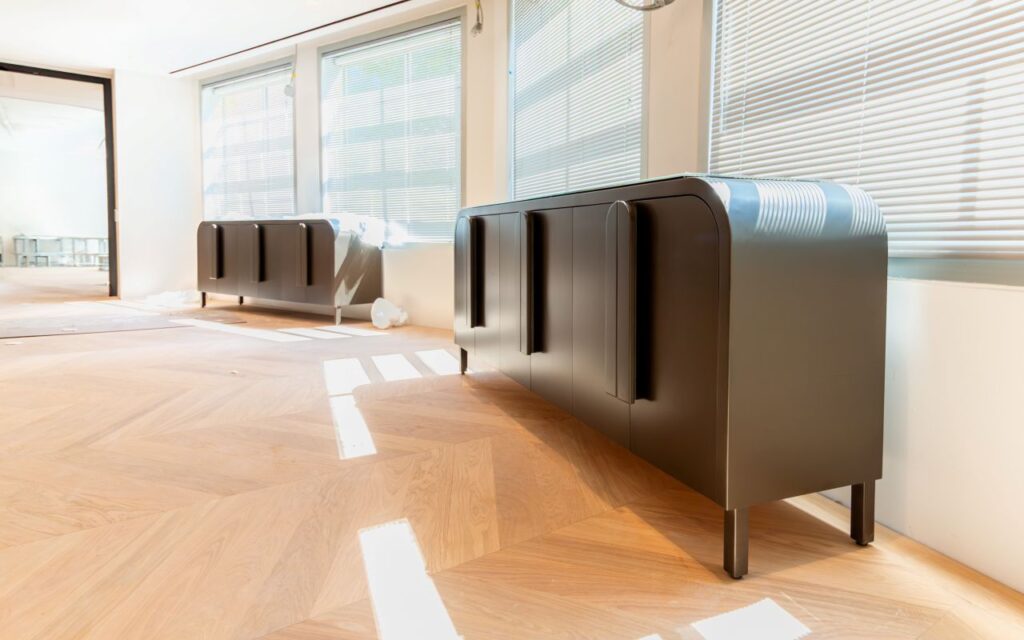 An angled wide shot of the minimalist office interior, showing two identical dark-metal sideboards with rounded edges and vertical pull handles. The units are placed against a white wall under a series of windows with silver horizontal blinds. Sunlight streams through the blinds, creating sharp, rectangular highlights on the light-oak chevron floors. The far background shows an open doorway and a glimpse of an unfinished workspace.