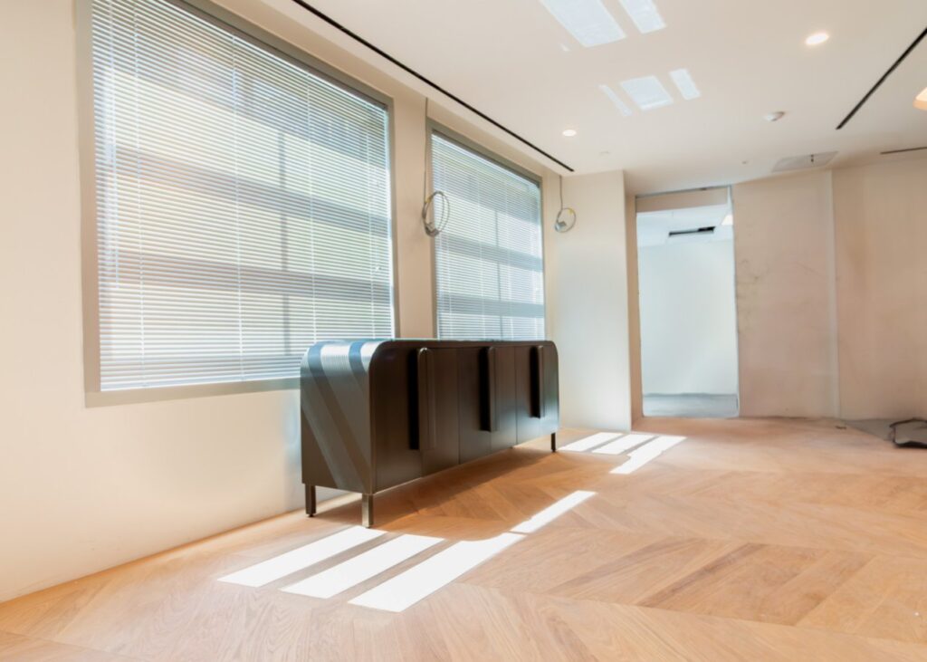 A bright, minimalist office interior featuring a sleek, dark-metal sideboard with curved edges and vertical door handles. The unit is positioned under two large windows with horizontal blinds, casting geometric shadows across a light-toned chevron-patterned wood floor. The room is in a state of completion or light construction, with clean white walls, recessed ceiling lights, and an open doorway leading to an adjacent white-walled space.