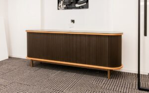 A high-angle, wide shot of the dark-wood slatted sideboard at 1100 NY Ave, emphasizing its elegant pill-shaped form and fine vertical ribbing. The unit is bordered by a light-oak top and bottom trim, supported by minimalist cylindrical legs in a muted bronze finish.