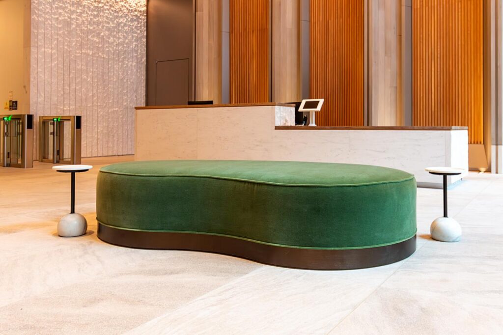 A modern office lobby featuring a large, kidney-shaped green velvet ottoman with a dark wood base. Two minimalist side tables with white marble bases flank the ottoman. In the background, there is a white marble reception desk and a warm, vertical-slatted wood wall.