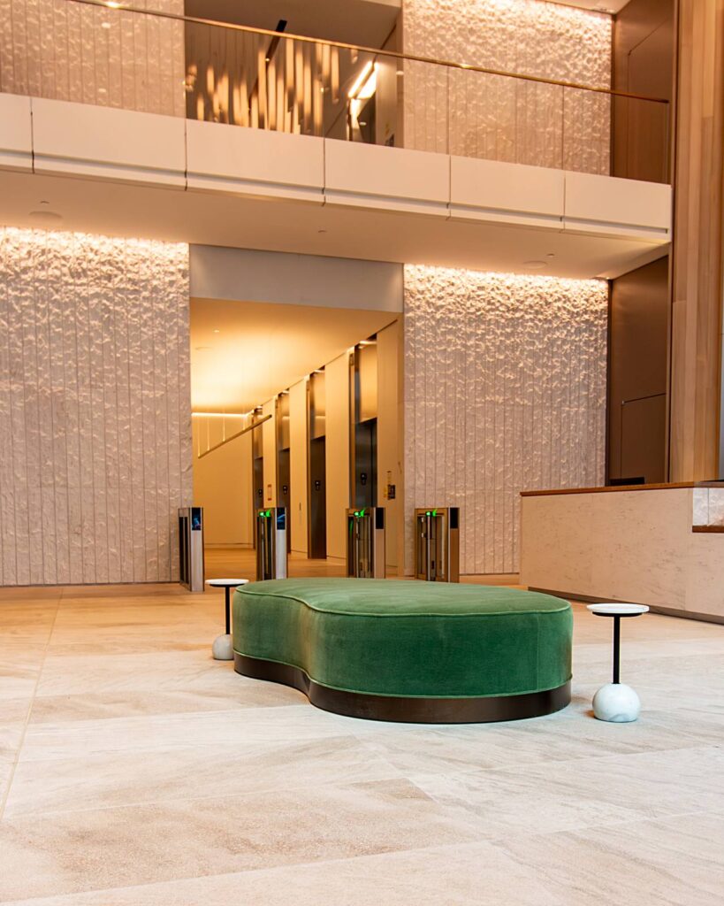 A wide-angle shot of a luxurious two-story building lobby, centered on the green velvet kidney-shaped ottoman and minimalist side tables from the previous view. The background reveals an elevator bank guarded by security turnstiles, set between walls of textured, backlit white stone and warm wood paneling. A glass-railed mezzanine overlooks the space from above.