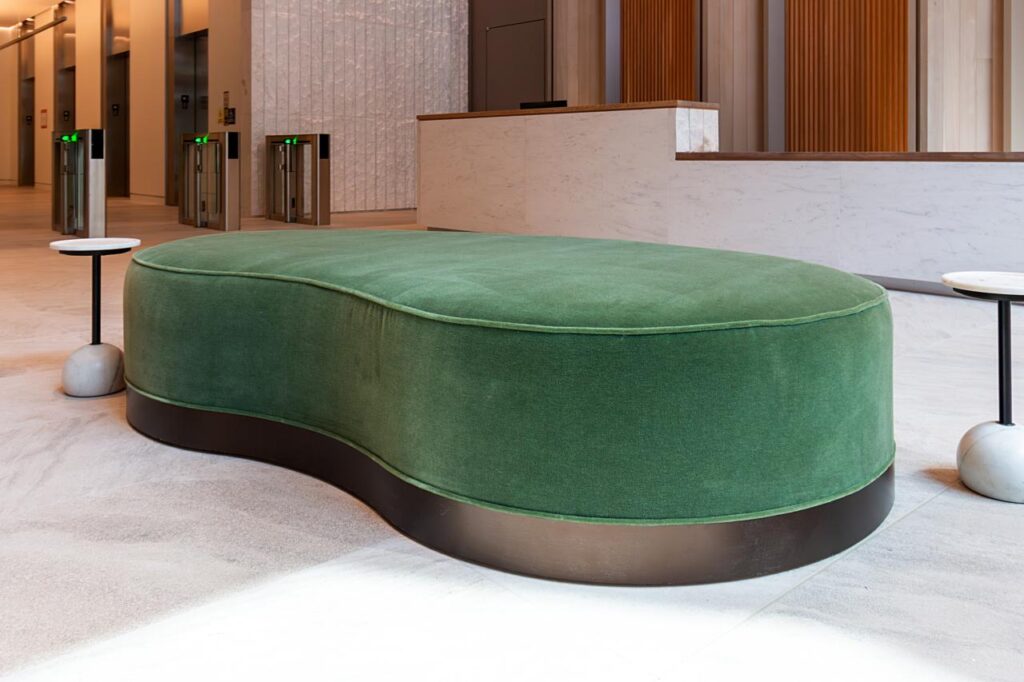 A close-up shot of the green velvet kidney-shaped ottoman, emphasizing its soft texture and dark, metallic-finish base. It is situated on a light-colored stone floor and flanked by two minimalist side tables with spherical marble bases. The background shows a partial view of the white marble reception desk and security turnstiles, maintaining a shallow depth of field.