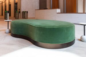 A close-up shot of the green velvet kidney-shaped ottoman, emphasizing its soft texture and dark, metallic-finish base. It is situated on a light-colored stone floor and flanked by two minimalist side tables with spherical marble bases. The background shows a partial view of the white marble reception desk and security turnstiles, maintaining a shallow depth of field.