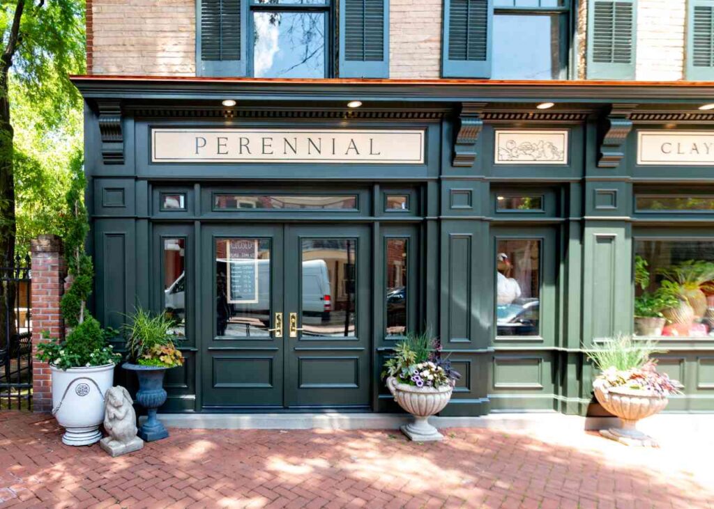 Street-level view of Perennial's charming storefront on a brick sidewalk, with dark sage green painted facade, decorative planters, and a small stone bunny statue flanking the entrance.