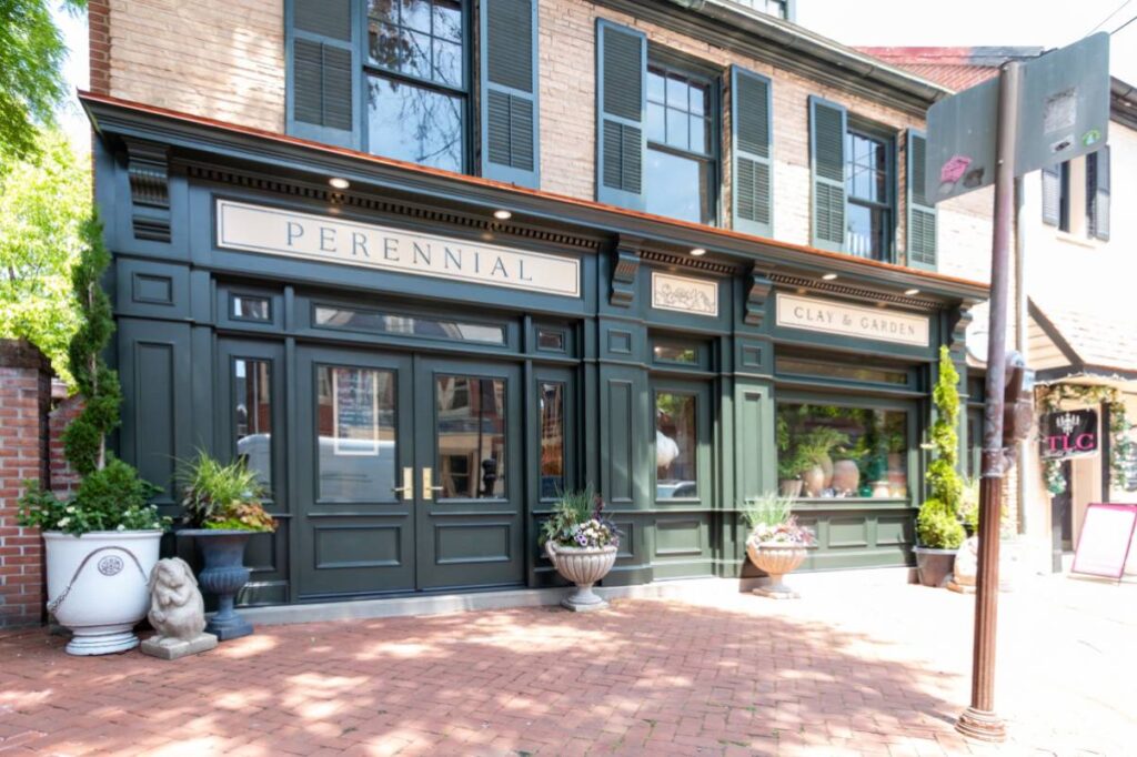 Short version: "Corner street view of Perennial and Clay & Garden storefronts sharing a dark sage green facade on a sunny brick sidewalk in a charming historic neighborhood