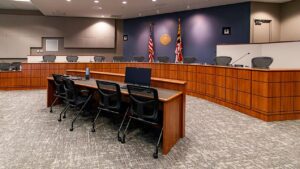 A government council chamber featuring a curved wood panel dais with microphones and name placards, an official seal, an American flag and a Maryland state flag on a navy blue accent wall, a clerk's desk with a laptop and black mesh chairs in the foreground, carpet flooring, and recessed ceiling lighting.