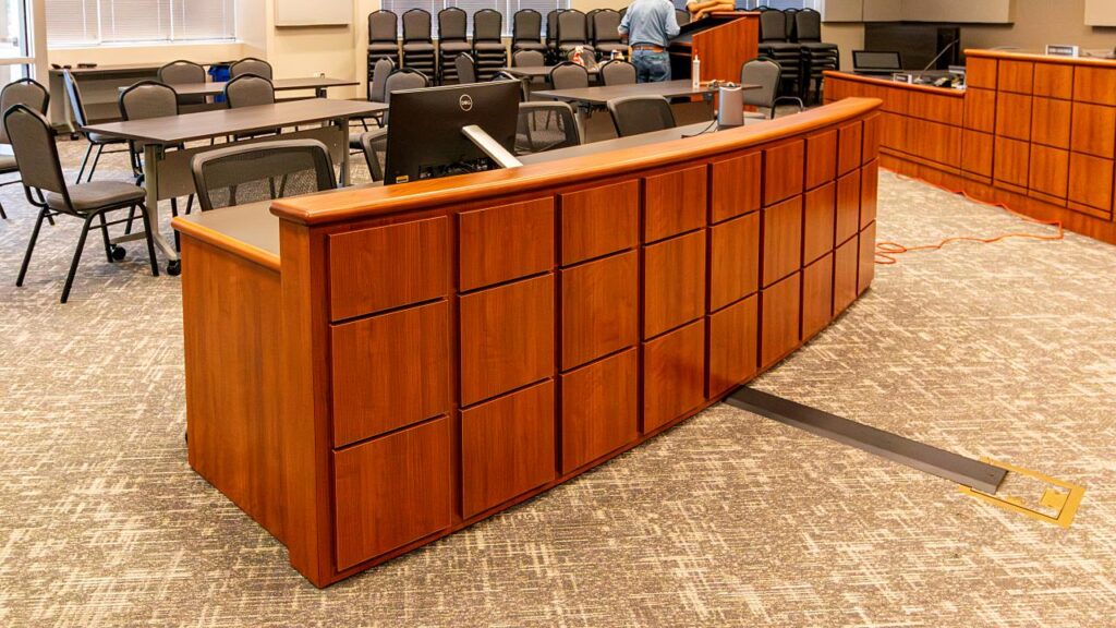 A curved wood clerk's desk with a grid-panel front in a warm cherry finish, topped with a Dell computer monitor, photographed during installation in a government council chamber with public seating, a dais, and a floor cable raceway visible in the foreground