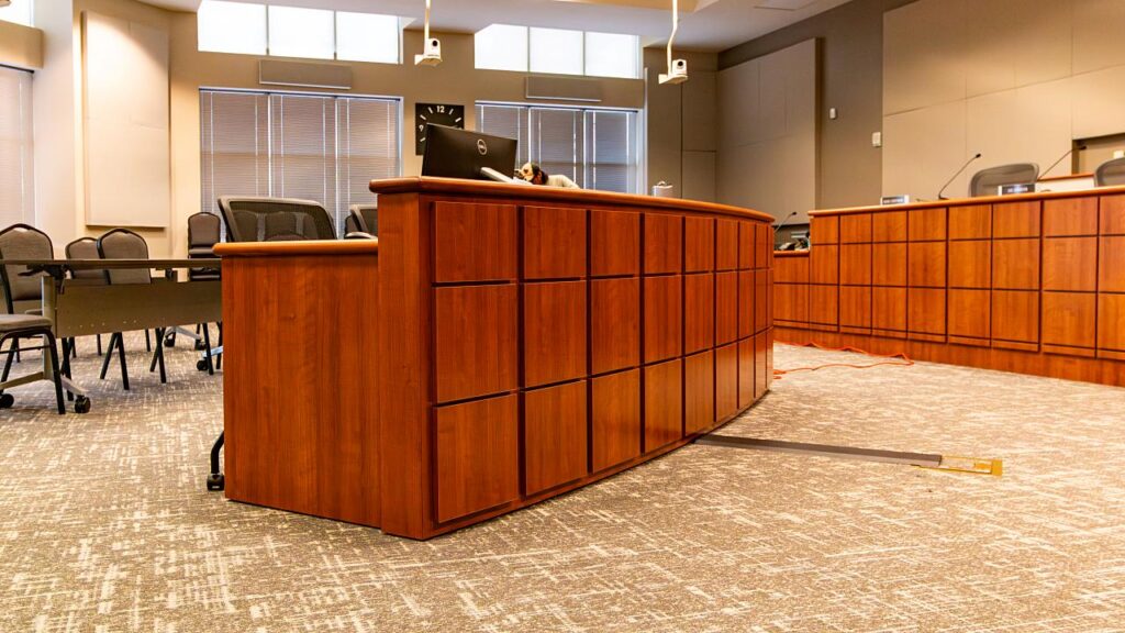 A curved cherry wood clerk's desk with a grid-panel front and computer monitor in a government council chamber, with the wood dais and council seating with microphones visible in the background, ceiling-mounted cameras, windows with blinds, public seating area to the left, and a floor cable raceway in the foreground.