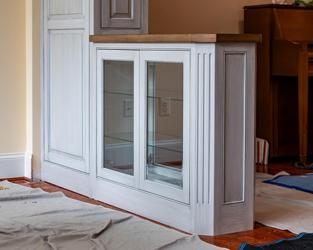 A built-in display cabinet with glass sliding doors and glass shelves, painted in a gray-white finish with raised panel details and a warm wood top, photographed during installation with drop cloths on the hardwood floor.