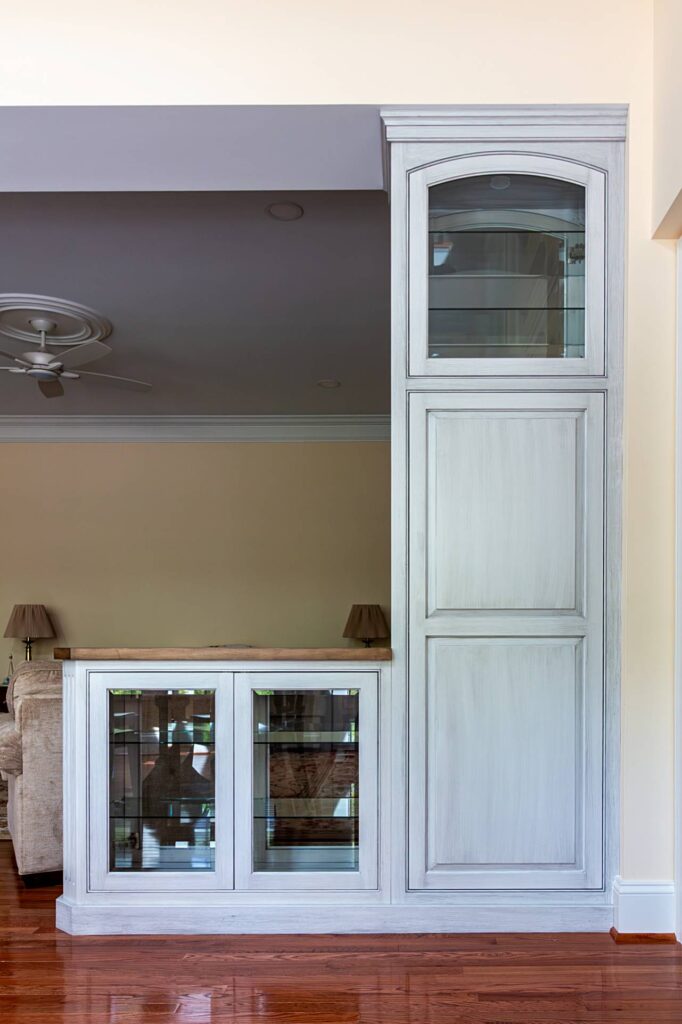 A tall built-in display cabinet with a gray-white distressed painted finish, featuring an arched glass upper door with glass shelves, two raised panel middle doors, and a lower glass-door display section with a wood countertop, installed on hardwood floors with a living room visible in the background.
