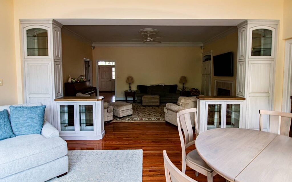 A wide doorway between a dining room and living room flanked by two matching tall built-in display cabinets with gray-white distressed painted finish, arched glass upper doors, raised panel middle sections, glass lower display cases, and wood countertops, with hardwood floors throughout and a living room with sofa, armchairs, ceiling fan, and wall-mounted TV visible beyond.