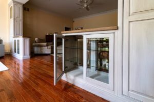 A close-up of a built-in low display cabinet with gray-white distressed painted finish, one glass door open revealing empty glass shelves inside, a wood countertop on top, set on polished hardwood floors with a living room and matching cabinetry visible in the background.