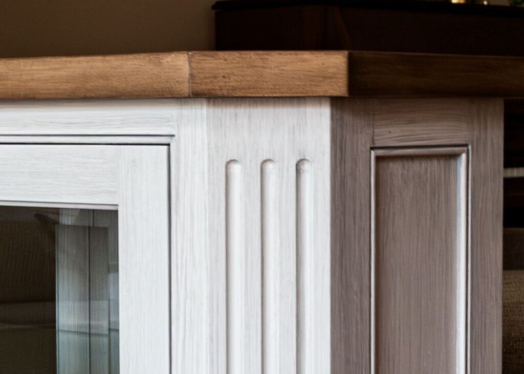 A close-up detail of a built-in cabinet corner showing a gray-white distressed painted finish with fluted pilaster detailing, a raised panel side, a glass door on the left, and a warm wood countertop on top.