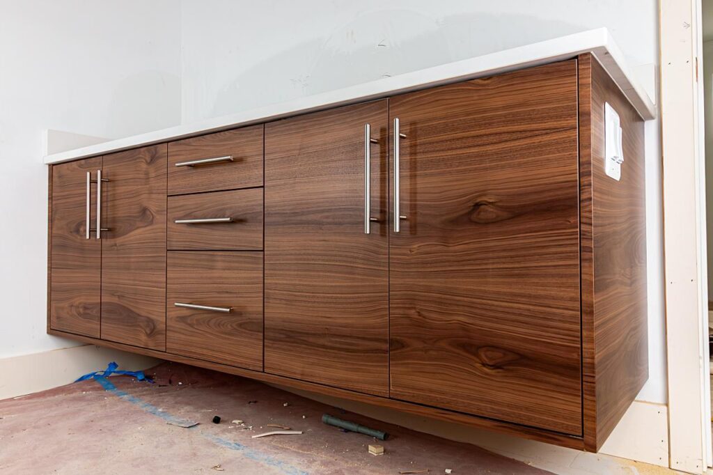 A floating walnut veneer bathroom vanity with bookmatched grain panels, three central drawers, two cabinet doors, brushed stainless steel bar pulls, and a white countertop, photographed during construction with debris on the floor.