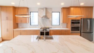 A completed modern kitchen with symmetrical light wood flat-panel cabinetry, a large white marble island with undermount sink in the foreground, matching marble slab backsplash behind a stainless steel wall-mounted range hood, a professional gas range, double wall oven, French door refrigerator, subway tile backsplash, two circular ring pendant lights, and hardwood floors.