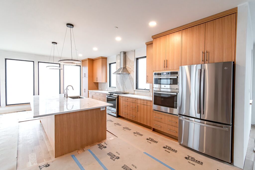 A modern kitchen under construction featuring light wood flat-panel cabinetry, a marble-topped island with undermount sink, stainless steel appliances including a gas range, double wall oven, and French door refrigerator, a stainless steel wall-mounted range hood with marble slab backsplash, a circular ring pendant light, large black-framed windows, and protective floor covering over hardwood floors.