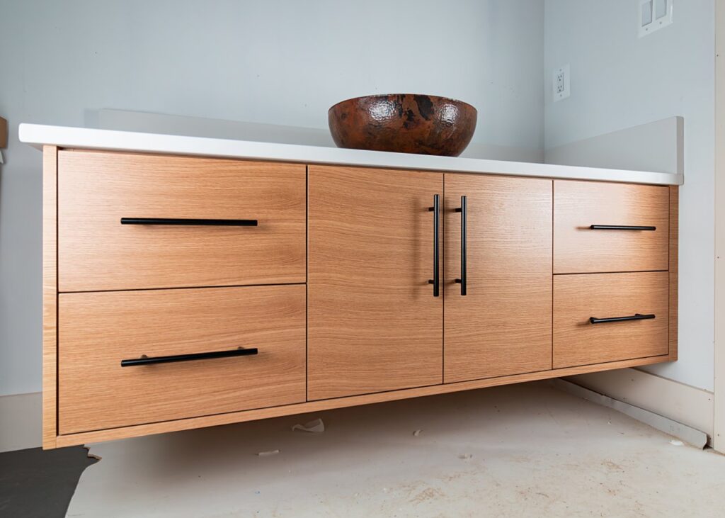 A floating bathroom vanity with light oak wood veneer panels, four drawers, and a center double-door cabinet with matte black bar pulls, topped with a white countertop and a hammered copper vessel sink bowl, photographed during construction.