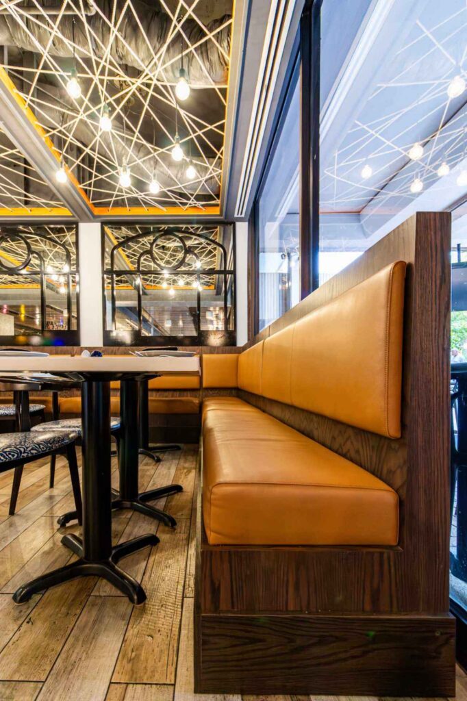 A low-angle view of the restaurant's tan leather banquette, showing the rich dark wood grain of the seating frame. The shot highlights an intricate ceiling installation of crisscrossing white ropes and hanging Edison bulbs. In the background, black-framed arched mirrors reflect the nautical decor, while the floor is made of rustic light-wood planks.