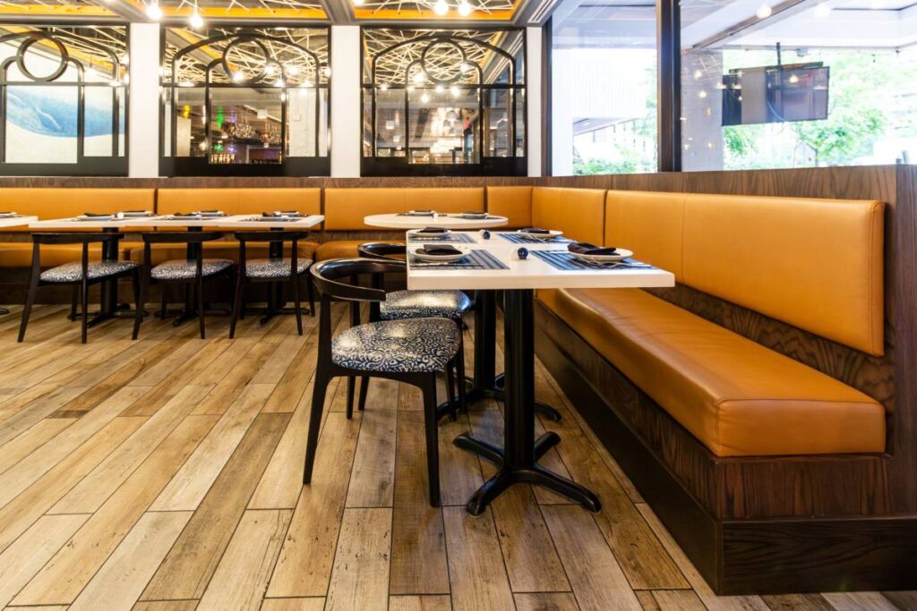 A wide-angle perspective of the nautical-themed dining area, showcasing the continuous L-shaped tan leather banquette and dark wood frame. The view highlights the contrast between the white tabletops and the patterned blue-and-white chair cushions, all set upon light-wood plank flooring. Large windows to the right bring in natural light, while the background features the signature arched mirrors and a glimpse of the blue whale mural.