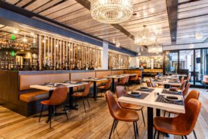 A wide-angle interior of the "All Set" restaurant dining room, featuring tan leather banquettes and cognac-colored chairs at white tables. The space is defined by an industrial ceiling with large crystal-style chandeliers and a unique partition made of vertically hanging ropes in varying shades of tan, white, and blue. The rustic wood-plank flooring and large windows create a bright, airy coastal atmosphere.