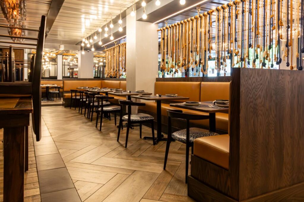 A warm, atmospheric shot of the "All Set" dining room highlighting the chevron-patterned wood floors. The scene features dark wood booths with tan leather upholstery and black dining chairs with blue-and-white patterned seats. In the background, a curtain of vertically hanging ropes with decorative knots serves as a room divider, complemented by a string of Edison bulb pendant lights that cast a soft glow across the industrial-style ceiling.