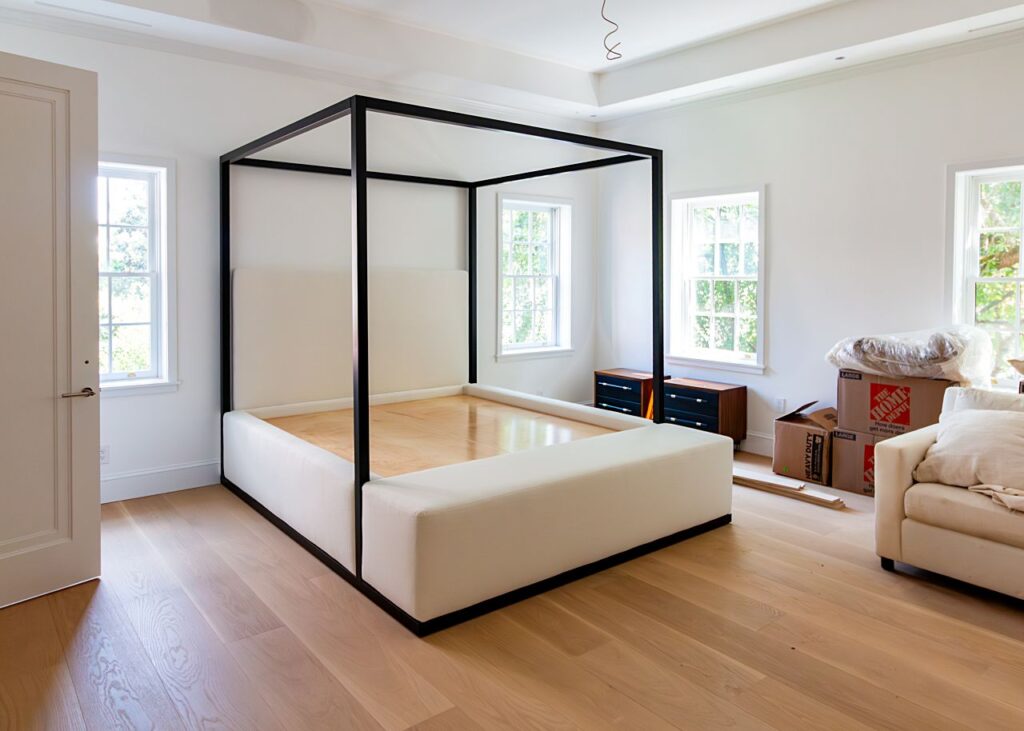 A modern four-poster canopy bed frame with a matte black metal square canopy structure, white upholstered headboard and platform base, without a mattress, in a bright bedroom with light hardwood floors, tray ceiling, multiple windows, moving boxes, a walnut nightstand, and a cream sofa visible during home installation