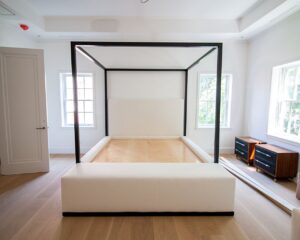 A front view of a modern four-poster canopy bed with a matte black square metal frame, white upholstered headboard and platform base without a mattress, in a bright bedroom with light herringbone hardwood floors, tray ceiling, multiple windows, and a dark navy and walnut dresser visible to the right.