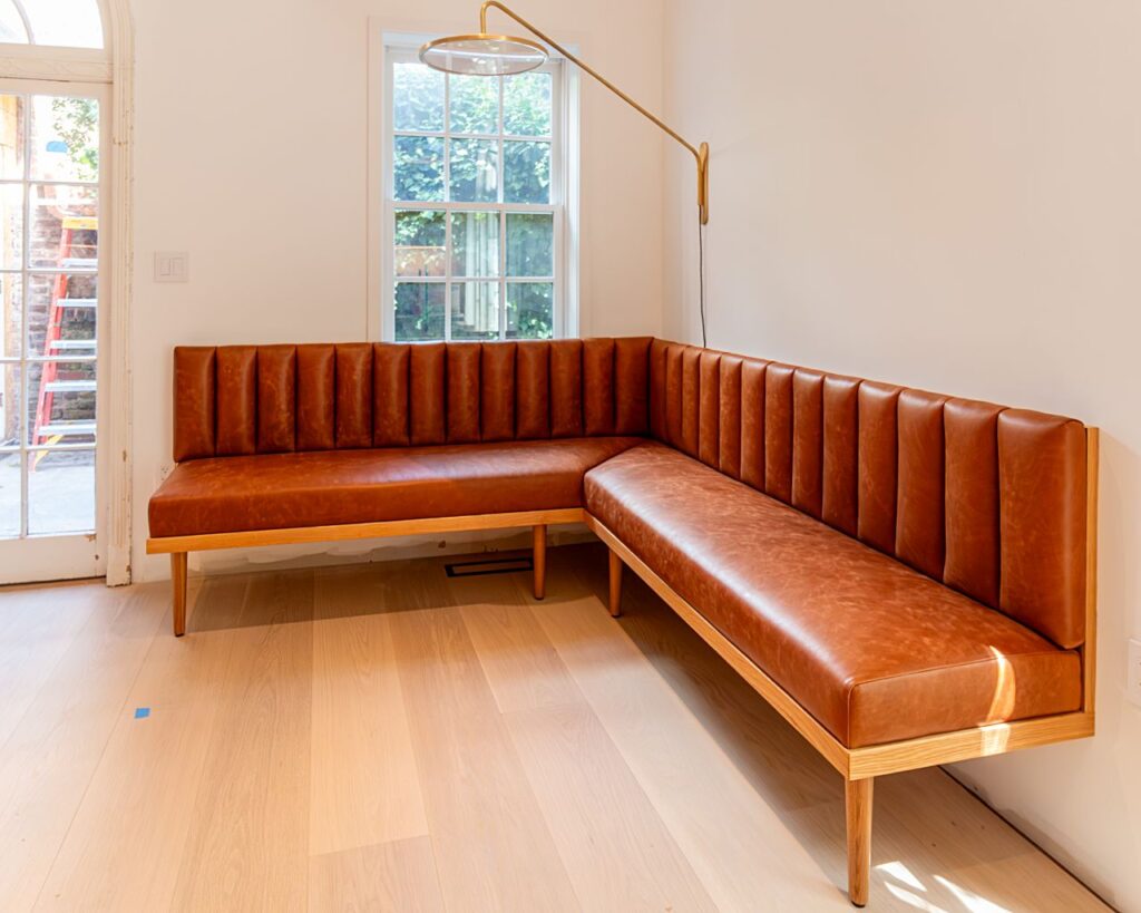 An L-shaped corner banquette with channel-tufted cognac leather upholstery, a light wood frame and tapered legs, positioned against two walls with a window, an articulating brass wall sconce above, light hardwood floors, and a French door with construction scaffolding visible outside.