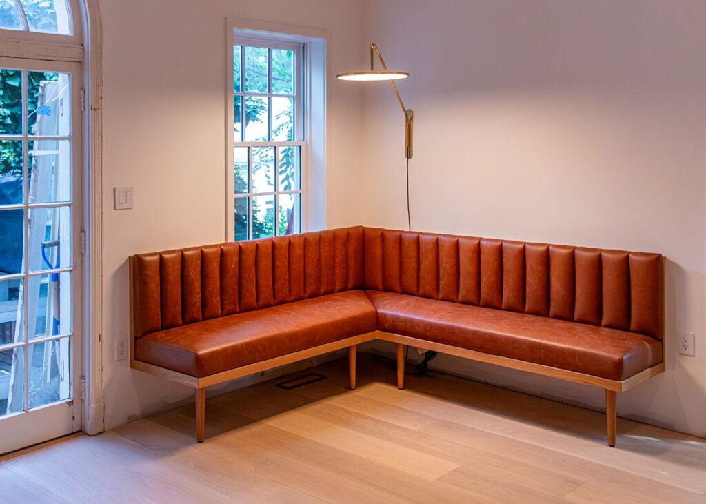 An L-shaped corner banquette with channel-tufted cognac leather upholstery and a light wood frame with tapered legs, wall-mounted against two white walls with a window, an illuminated articulating brass wall sconce above, light hardwood floors, and a French door with construction scaffolding visible outside
