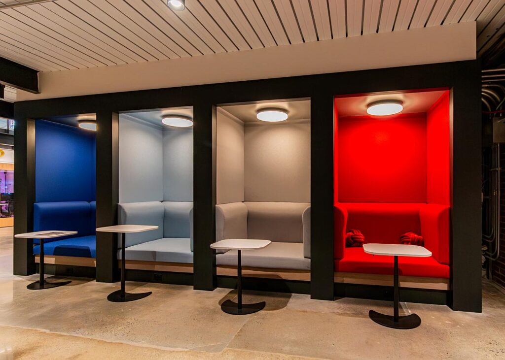 A row of four vibrant, color-coded semi-private seating alcoves set into a black wall. From left to right, the booths are royal blue, light blue, gray, and bright red. Each alcove features a matching upholstered bench, an overhead circular light, and a small white teardrop-shaped pedestal table on a black base.