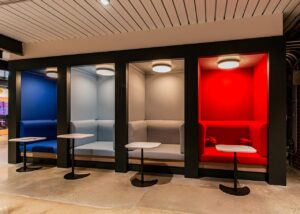 A row of four vibrant, color-coded semi-private seating alcoves set into a black wall. From left to right, the booths are royal blue, light blue, gray, and bright red. Each alcove features a matching upholstered bench, an overhead circular light, and a small white teardrop-shaped pedestal table on a black base.