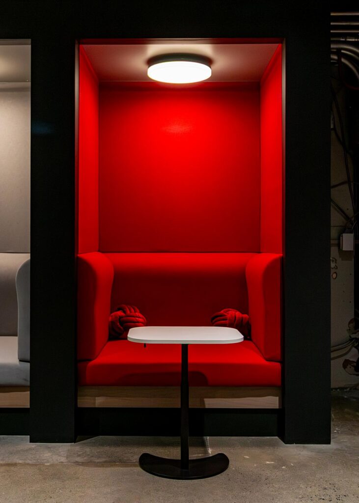 A close-up shot of a single, vibrant red semi-private booth. The alcove features a high-back upholstered red bench with two unique, knotted red pillows, a small white teardrop-shaped table, and a circular overhead light that casts a warm glow against the red back wall. The booth is framed by thick black partitions on a polished concrete floor.