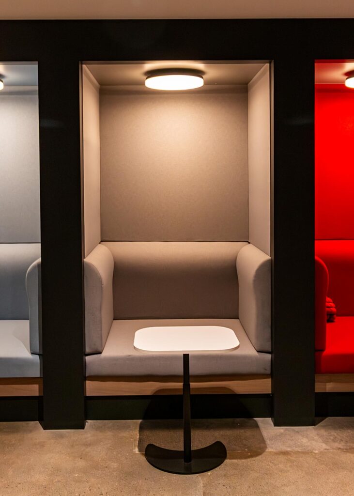 A close-up shot of a minimalist, light-gray semi-private seating booth. The alcove is framed by thick black walls and features an upholstered gray bench seat and backrest, a white teardrop-shaped pedestal table, and a warm circular overhead light. Part of a light-blue booth is visible to the left and a red booth to the right, all set on a polished concrete floor.