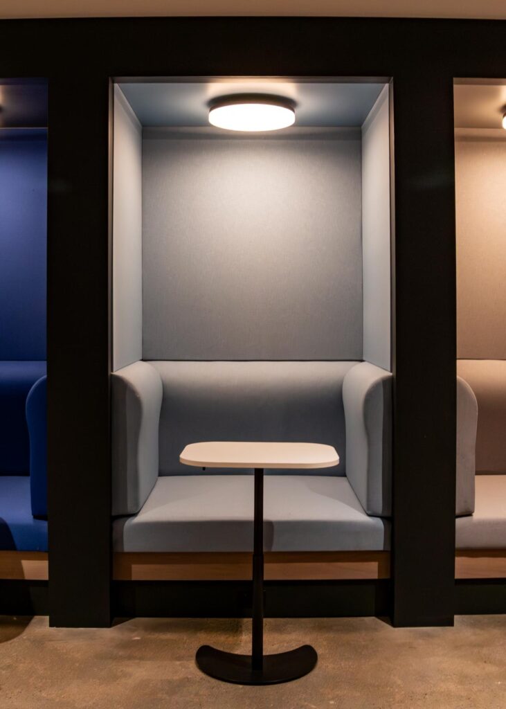 A close-up shot of a light-blue semi-private seating booth. The alcove is framed by thick black walls and features a matching light-blue upholstered bench seat and high backrest. A small, white teardrop-shaped pedestal table sits in front of the bench, and a circular overhead light provides a soft glow. Portions of a royal blue booth to the left and a gray booth to the right are visible on a polished concrete floor.