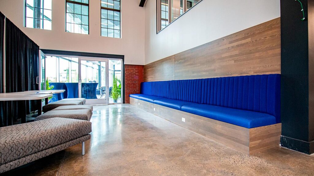 A wide-angle view of a high-ceilinged industrial space featuring a long, royal blue channel-tufted banquette built into a wood-paneled wall. Across from the bench are three textured gray ottomans on metal legs and a white table. The room features polished concrete floors, large multi-pane windows, and a glass door leading to an outdoor patio.