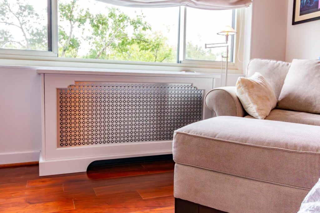 A white, custom-built radiator cover with a decorative metallic lattice grille situated under a window in a bright living room. The cover features a scalloped architectural cutout and is flanked by a tan-and-white checkered armchair and ottoman on polished cherry wood floors.