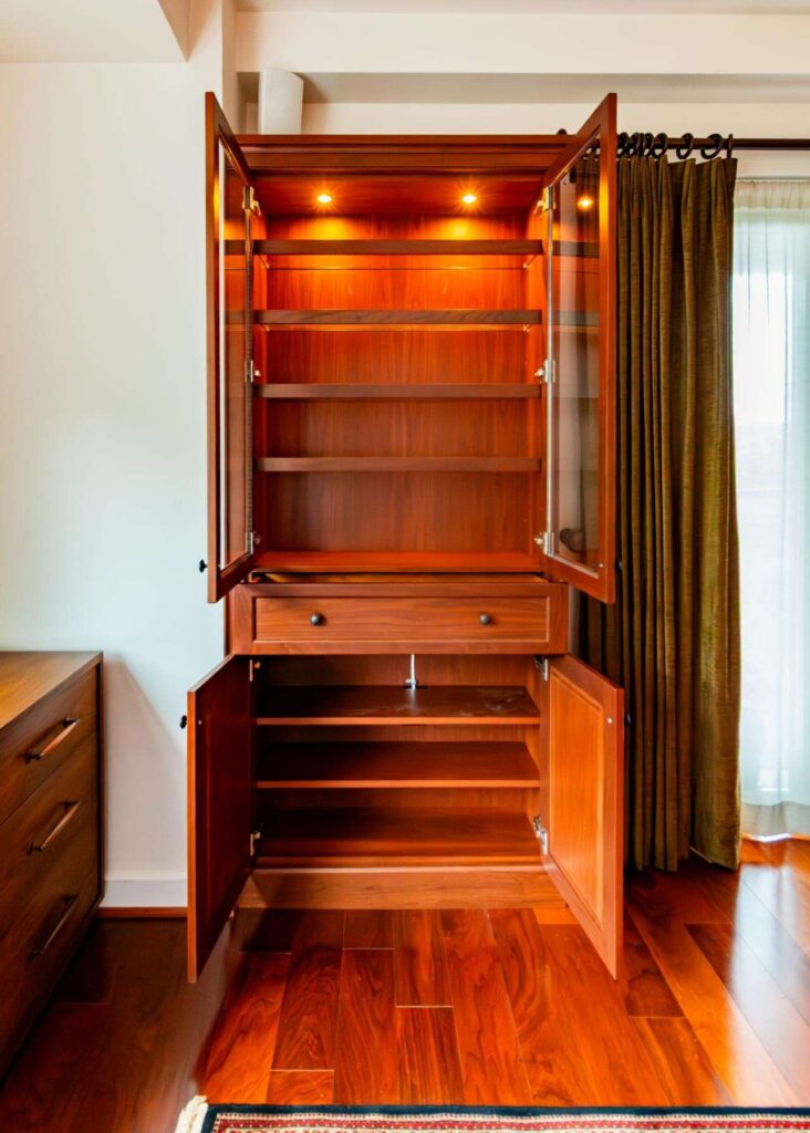 A full-frontal view of the dark wood hutch from the previous image, now with all doors and its central drawer open to showcase the interior storage. The upper glass-fronted section reveals several shelves illuminated by two warm puck lights at the top, while the lower solid-door section shows additional wooden shelving. The unit stands on polished cherry floors beside a heavy brown curtain.