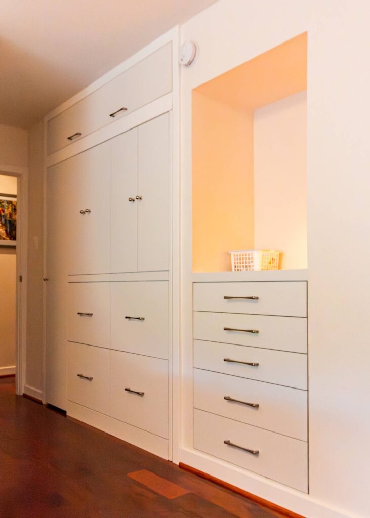 A frontal view of the custom white hallway built-ins, showing the unit fully closed. To the left, large cabinets and wide drawers feature small round knobs and brushed-metal handles. To the right, a vertical stack of five drawers sits beneath a brightly illuminated recessed alcove. The entire unit is integrated into the wall and set against dark cherry wood flooring.