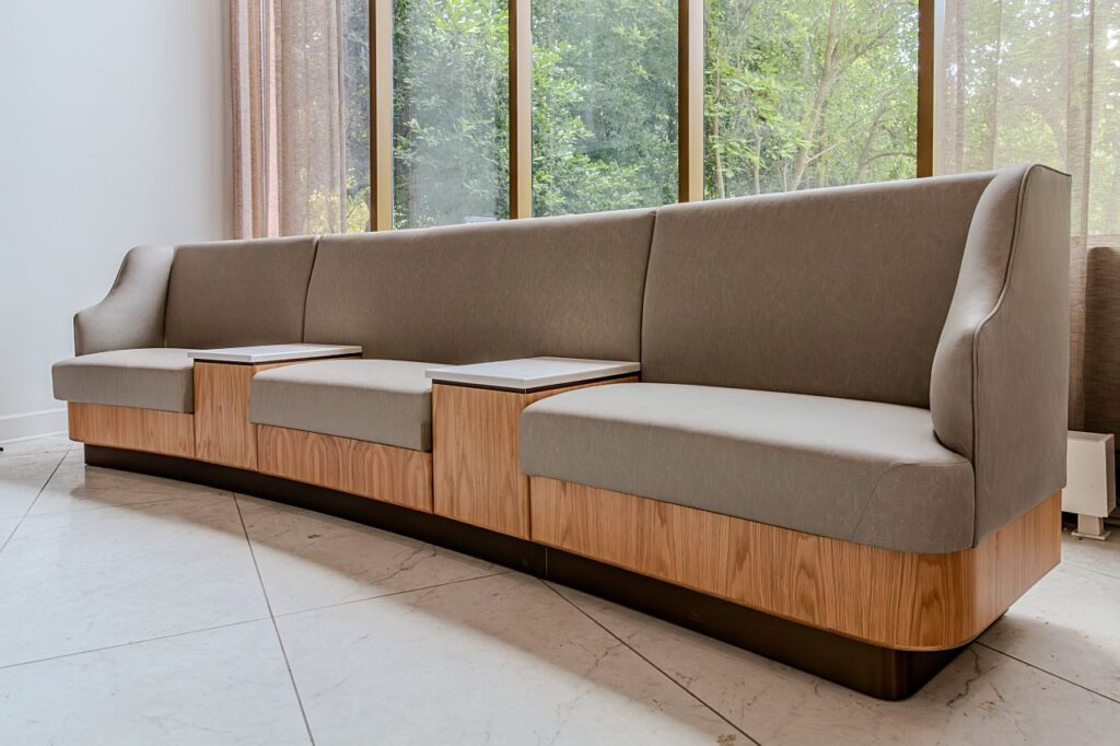 A custom modular banquette seating unit with high backs upholstered in taupe fabric, light wood grain panel bases, integrated side tables with stone tops, on a dark plinth base, positioned against floor-to-ceiling windows with sheer curtains and a garden view beyond.