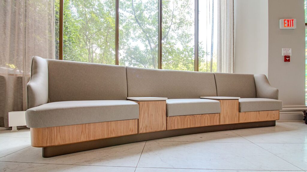 A custom modular banquette seating unit with high backs upholstered in taupe fabric, light oak wood grain panel bases, integrated white stone side tables, on a dark plinth base, positioned against floor-to-ceiling windows with sheer curtains and a tree view, with an exit sign and fire alarm visible on the wall.