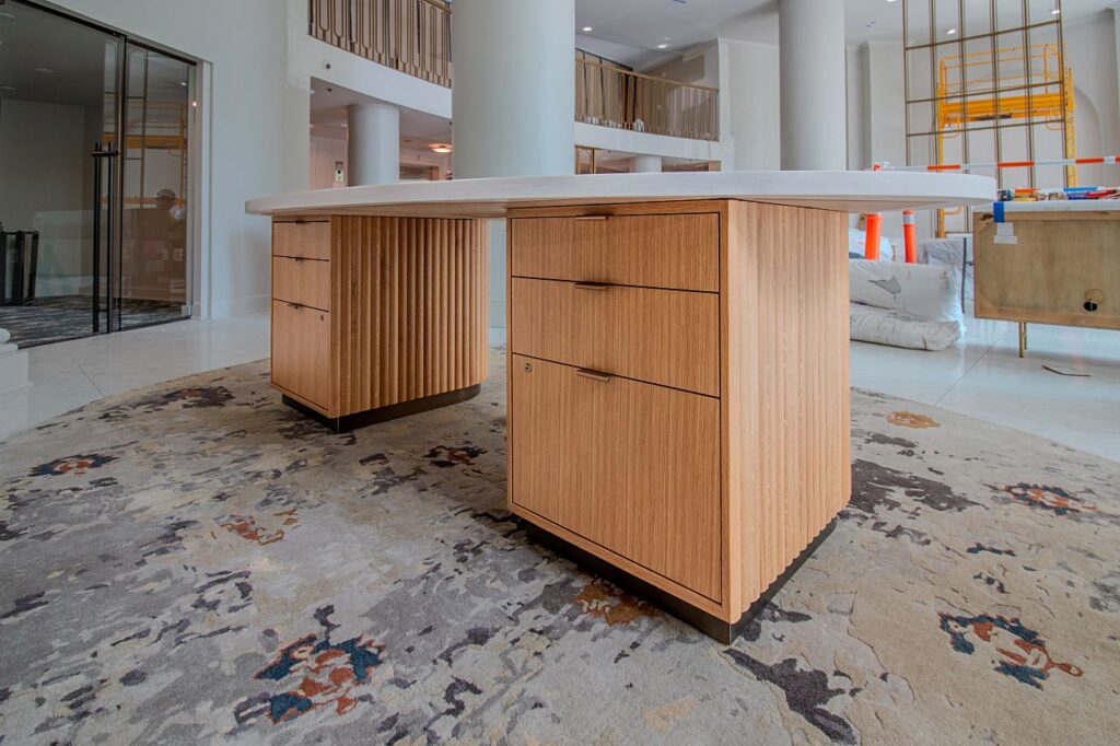 A custom executive desk with a curved white stone top, light oak wood pedestals featuring fluted panel and flat drawer fronts with dark metal pulls, positioned on a decorative area rug in a grand hotel or commercial lobby under construction, with tall columns, a mezzanine level, and construction scaffolding visible in the background.