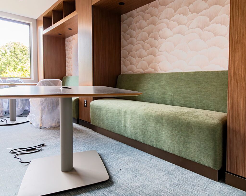 A built-in banquette booth with green upholstered seating set within a warm wood-paneled niche featuring floral patterned wallpaper, open shelving above, and a power outlet on the side panel, paired with a pedestal table in the foreground, photographed during commercial interior construction.