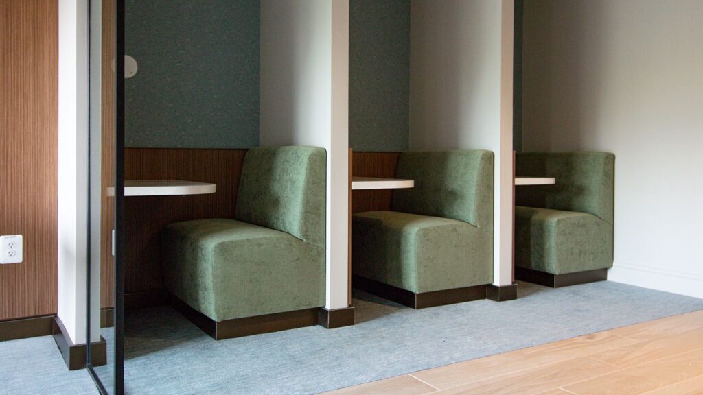 Three individual built-in booth seating alcoves with green upholstered armless chairs, small white side tables, dark wood wainscoting, teal acoustic panel backs, and dark base plinths, separated by white columns on a gray carpet, viewed through a glass partition in a commercial interior.