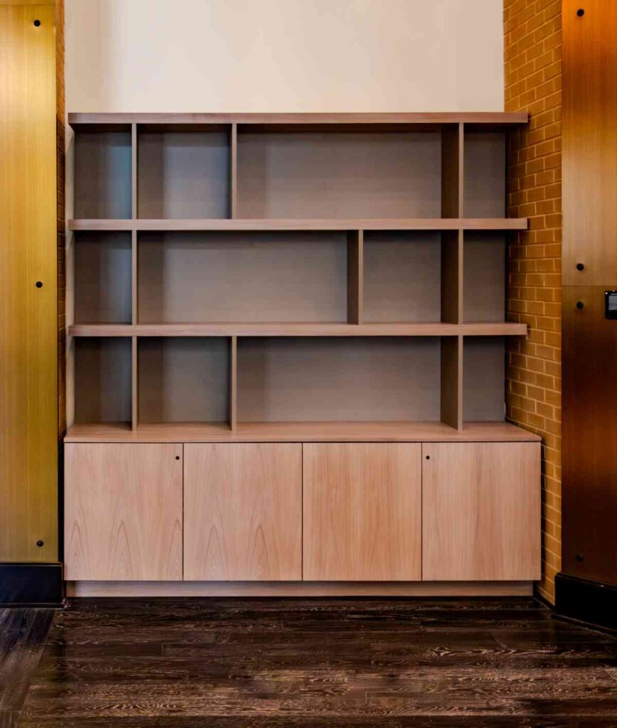 A built-in light wood shelving unit with three open display shelves and four lower cabinet doors, set against a cream wall flanked by orange brick and warm wood paneling