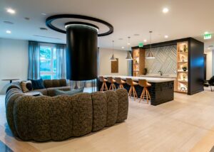 A modern residential amenity lounge featuring a curved modular sectional sofa in textured dark fabric surrounding a cylindrical black column with a circular LED ceiling feature, and in the background a bar area with a dark glazed brick tile island, white countertop, tan bar stools, brushed nickel pendant lights, lit arched display shelving, and a geometric patterned accent wall.