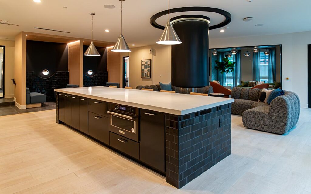 A large commercial kitchen island with a white countertop, black matte cabinetry, a built-in stainless steel speed oven, and a dark glazed brick tile end panel, with three brushed nickel cone pendant lights above, a cylindrical black column with circular LED ceiling feature nearby, and a lounge seating area with modular sofas and large windows visible in the background.