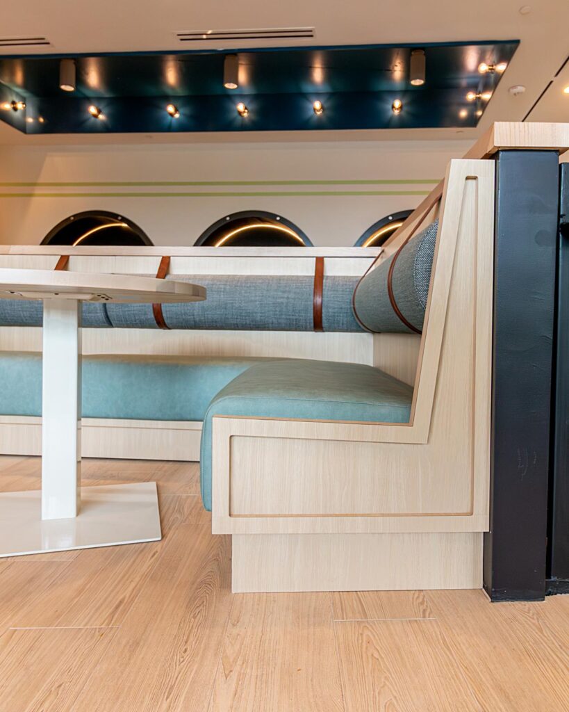 A detailed low-angle perspective of a light-wood-paneled booth with light-green leather and textured gray fabric seats, light wood table and white square base, set against a backdrop of arched wall accents with yellow LED lighting.