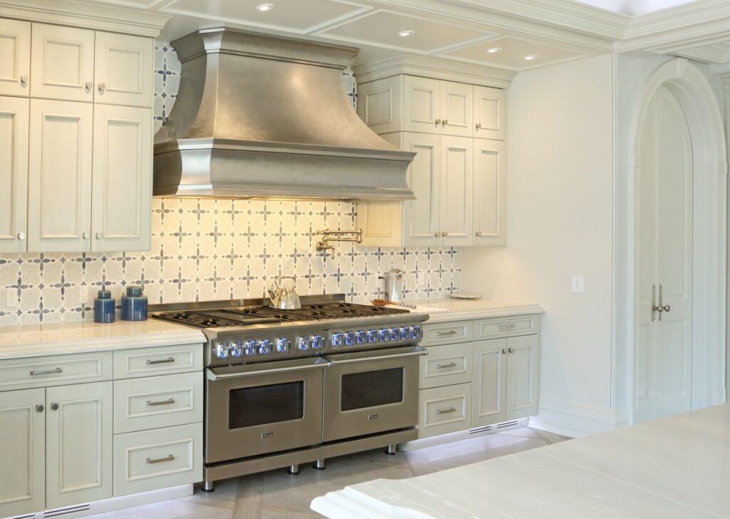 high-end kitchen featuring a professional-grade stainless steel double oven and range with blue LED-lit knobs. A large, matching brushed metal vent hood is mounted against a white and blue quatrefoil tile backsplash.